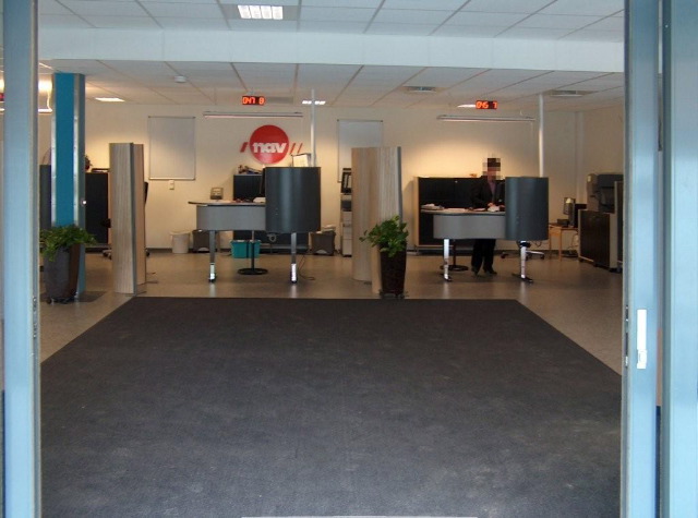 the front reception area with a dark grey rug providing a color contrast and leading visitors toward the backlit reception area with the red NAV logo visible from the entry.