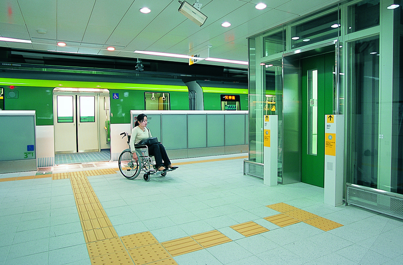 Woman in wheelchair exits train onto brightly lit platform and heads to a large glass elevator.