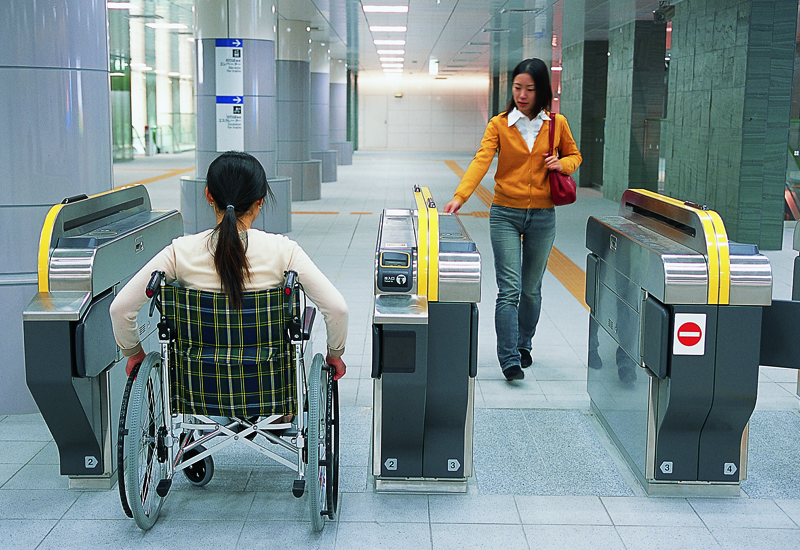 A woman in a wheelchair rolls through a wide fare gate as a woman walks through in the other direction