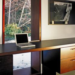 A large, deep, single pane window with a simple wooden desk table surface in front with a laptop on it.