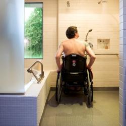 Siple in his wheelchair in the shower, shower has white wall tiles and light green floor tiles